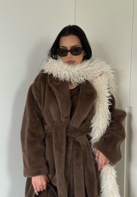 Manteau en fausse fourrure marron avec une taille ceinturée, assorti d'une écharpe blanche à poils longs. Lunettes de soleil foncées complètent le look. Texture lisse ; coupe oversize.