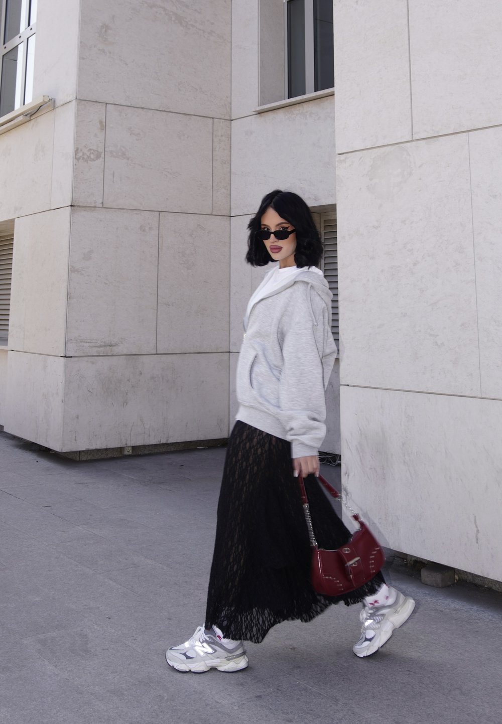 Grey oversized hoodie, white top, black lace skirt, silver trainers, and a small red handbag. Standing on a paved surface next to a building.