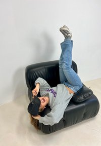 Black leather lounge chair with rounded edges and soft upholstery. An individual strikes a playful pose while wearing a grey sweatshirt and jeans.