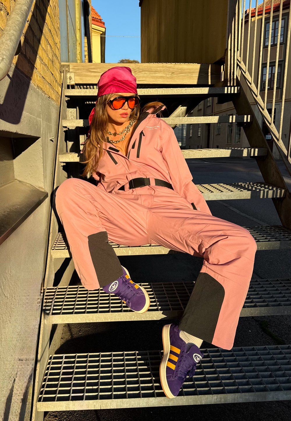 Frau in pinkem Overall, rotem Kopftuch, großen orangefarbigen Sonnenbrillen und lila Turnschuhen, die in der Sonne auf metallischen Außentreppen sitzt.