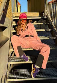 Frau in pinkem Overall, rotem Kopftuch, großen orangefarbigen Sonnenbrillen und lila Turnschuhen, die in der Sonne auf metallischen Außentreppen sitzt.