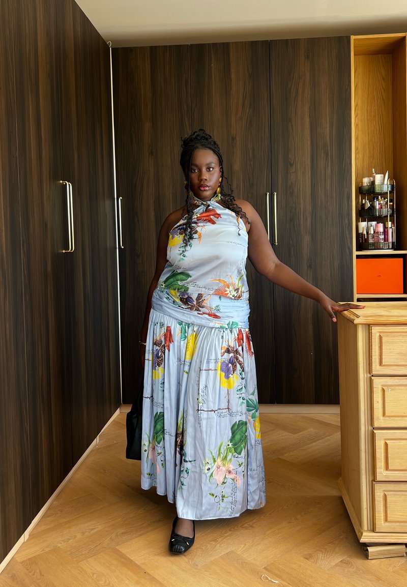 Femme aux cheveux tressés portant une robe à fleurs se tient à l'intérieur, s'appuyant sur une commode en bois, avec des armoires en bois foncé en arrière-plan.