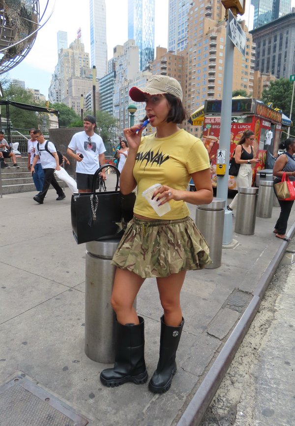 Vrouw in een gele shirt en camouflage rok, met zwarte laarzen en een pet, snackend bij een druk straatvoedselkraampje in New York City, omringd door wolkenkrabbers.