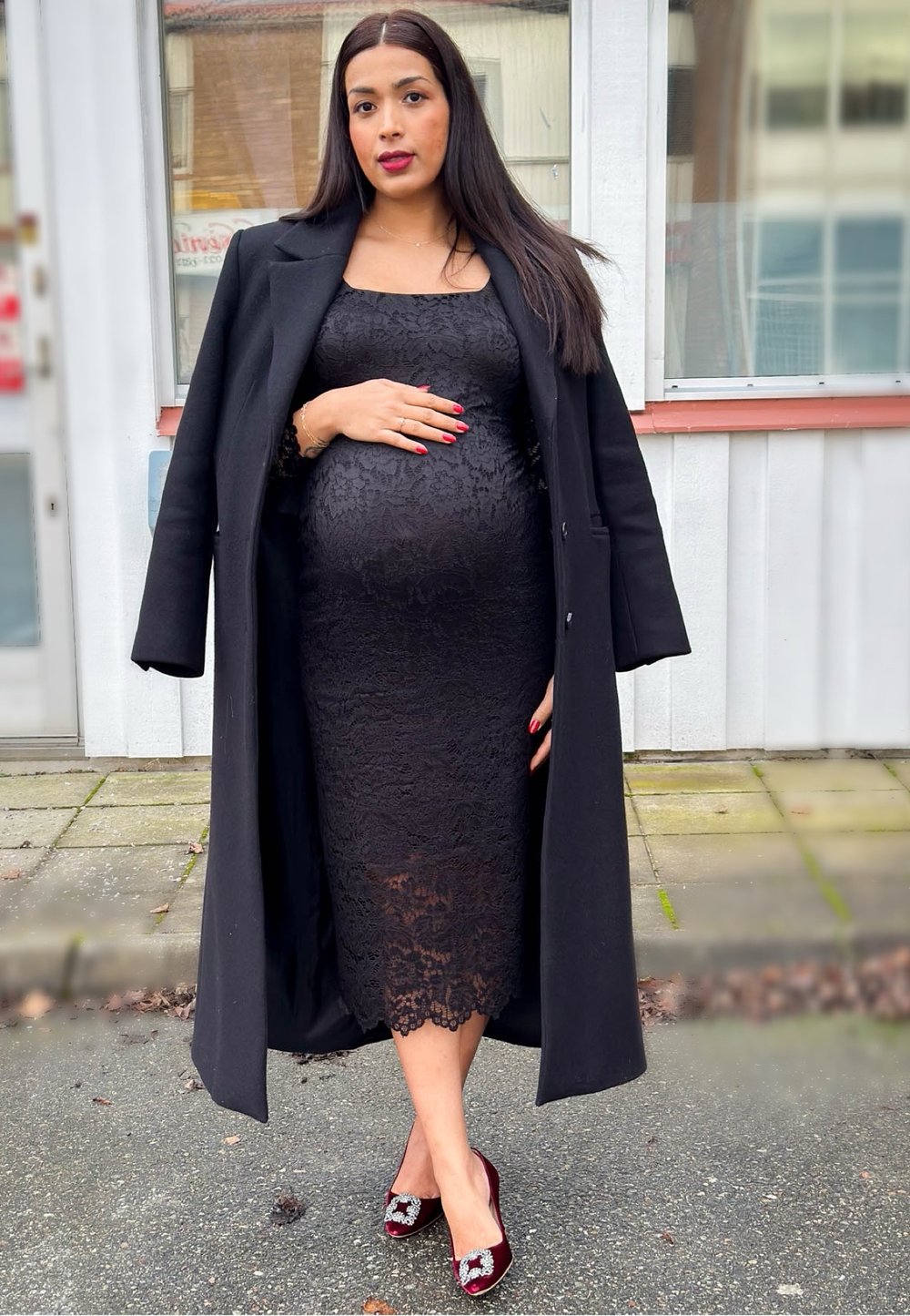 Abito in pizzo nero con silhouette aderente, cappotto nero lungo e décolleté bordeaux con dettagli decorativi. Capelli lisci e unghie rosse.