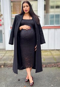 Abito in pizzo nero con silhouette aderente, cappotto nero lungo e décolleté bordeaux con dettagli decorativi. Capelli lisci e unghie rosse.