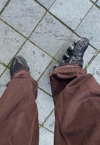 Jambes en pantalon marron et chaussures de sport noires posées sur un pavé en pierre grise avec de l'herbe poussant dans les interstices.