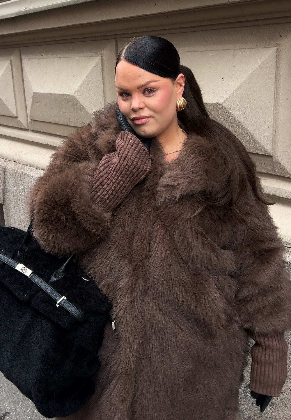 Brun fuskpälsrock med vid krage, ribbade bruna ärmar och svart handväska med silverdetaljer. Slätt hår stylat bakåt.