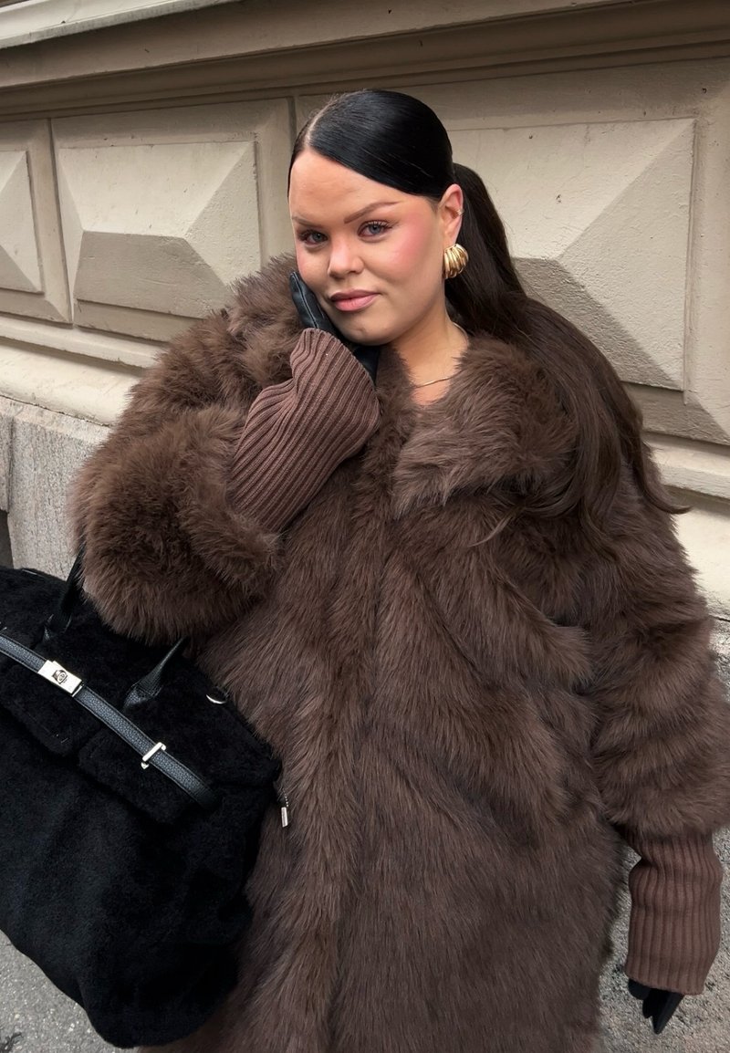 Cappotto in pelliccia sintetica marrone con ampio colletto, maniche marroni a coste e borsa nera con dettagli in argento. Capelli lisci pettinati all'indietro.