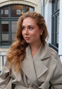 Beige-sävyinen trench-coat, jossa on suuri kaulus ja vyötärölle sidottu nauha. Hiukset on muotoiltu pehmeille aalloille, ja asusteina on kultainen klipsi ja korvakorut.