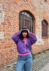 Jeune femme en veste violette et jeans ajustant un bonnet en tricot noir et beige devant un bâtiment en briques avec des fenêtres en arc.