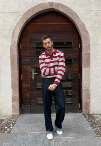 Punaste ja valgete triipudega pika varrukaga polo-särk, tumelavad sinised teksad ja valged tossud, seisab puidust ukse ees, millel on kaarjas raam.