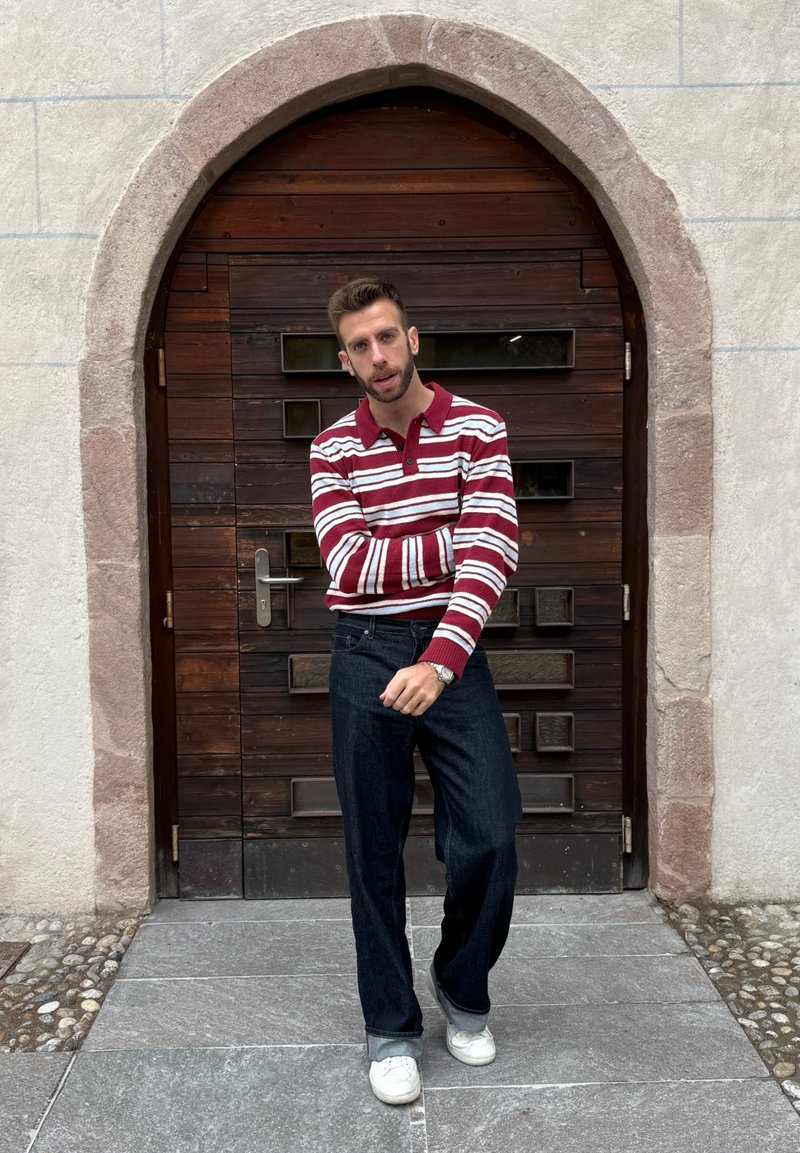 Maglietta polo a maniche lunghe a righe rosse e bianche, jeans blu scuro e scarpe da ginnastica bianche, in piedi davanti a una porta di legno con una cornice ad arco.