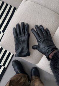 Gants en cuir noir sur un tissu beige texturé, un gant porté sur la main droite, personne portant des bottes noires et un pantalon marron visible.