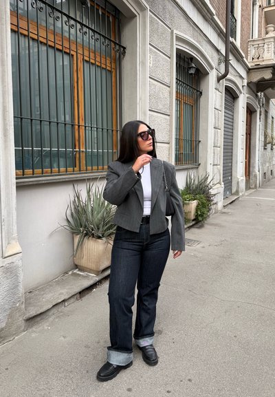 Blazer gris ajustado sobre una camisa blanca, combinado con jeans oscuros con mangas enrolladas. Calzado negro, gafas de sol de gran tamaño y fondo gris con maceta.