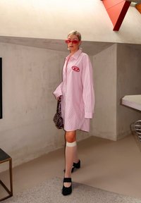 Överdimensionerad rosa randig skjorta med en ficklogotyp, kombinerad med svarta klackskor med blockklack och genomskinliga vita knähöga strumpor.