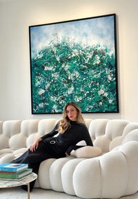 Γυναίκα με μαύρο φόρεμα ξαπλωμένη σε λευκό υφασμάτινο καναπέ κάτω από έναν μεγάλο πράσινο και μπλε αφηρημένο πίνακα, με βιβλία σε μαρμάρινο τραπέζι κοντά της.