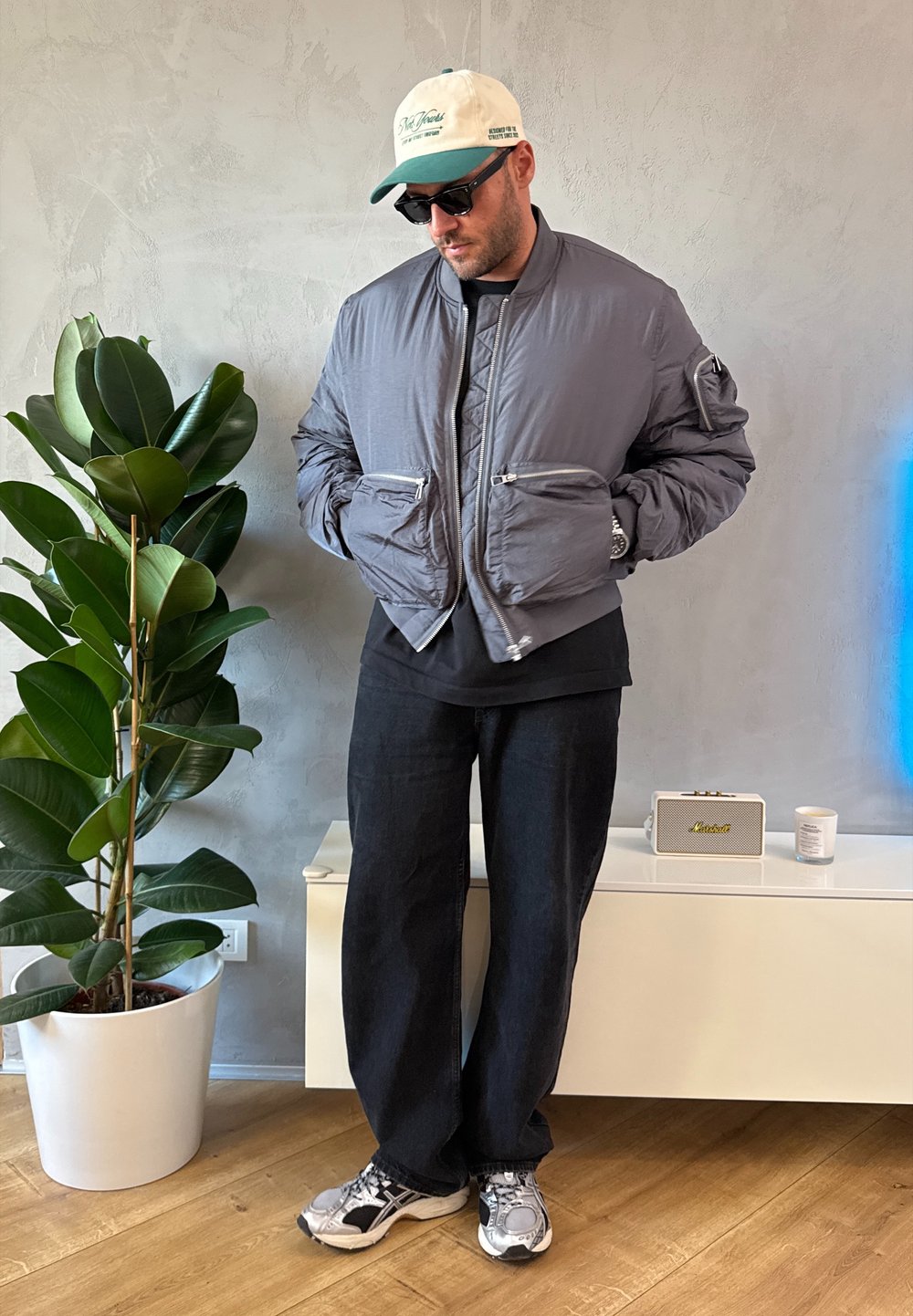 Giacca bomber grigia con collo a costine e molteplici tasche con cerniera, abbinata a pantaloni neri ampi e scarpe da ginnastica grigie. Ambiente interno con piante.