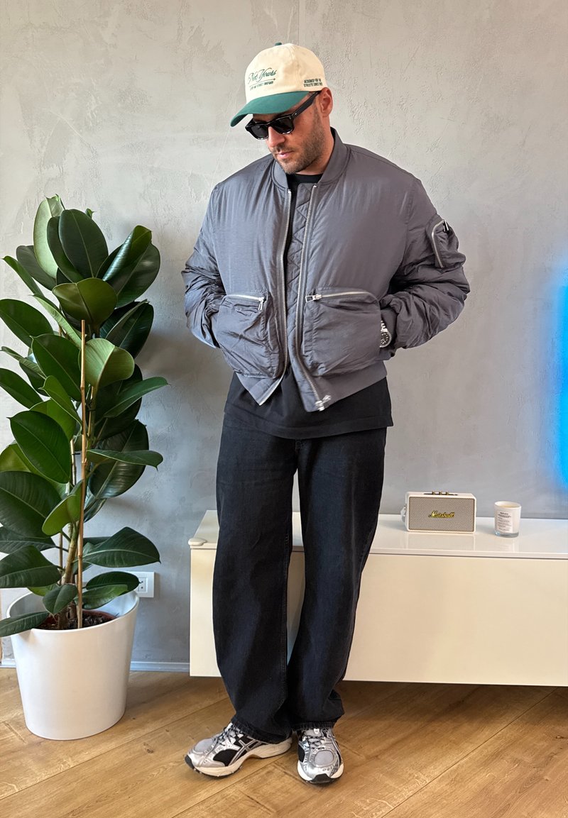 Graue Bomberjacke mit gerippter Kragenpartie und mehreren Reißverschlusstaschen, kombiniert mit schwarzen, locker sitzenden Hosen und grauen Turnschuhen. Innenraum mit Pflanzen.