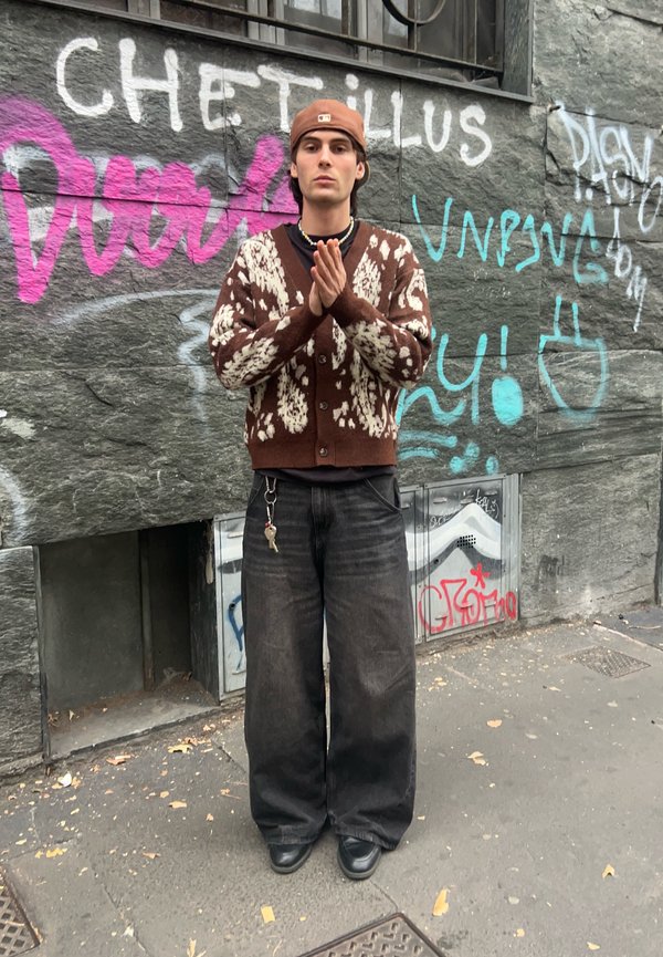 Barna kötött kardigán fehér mintákkal, bő fekete farmernadrág, fekete cipők és egy barna baseballsapka. Graffiti fal előtt áll.