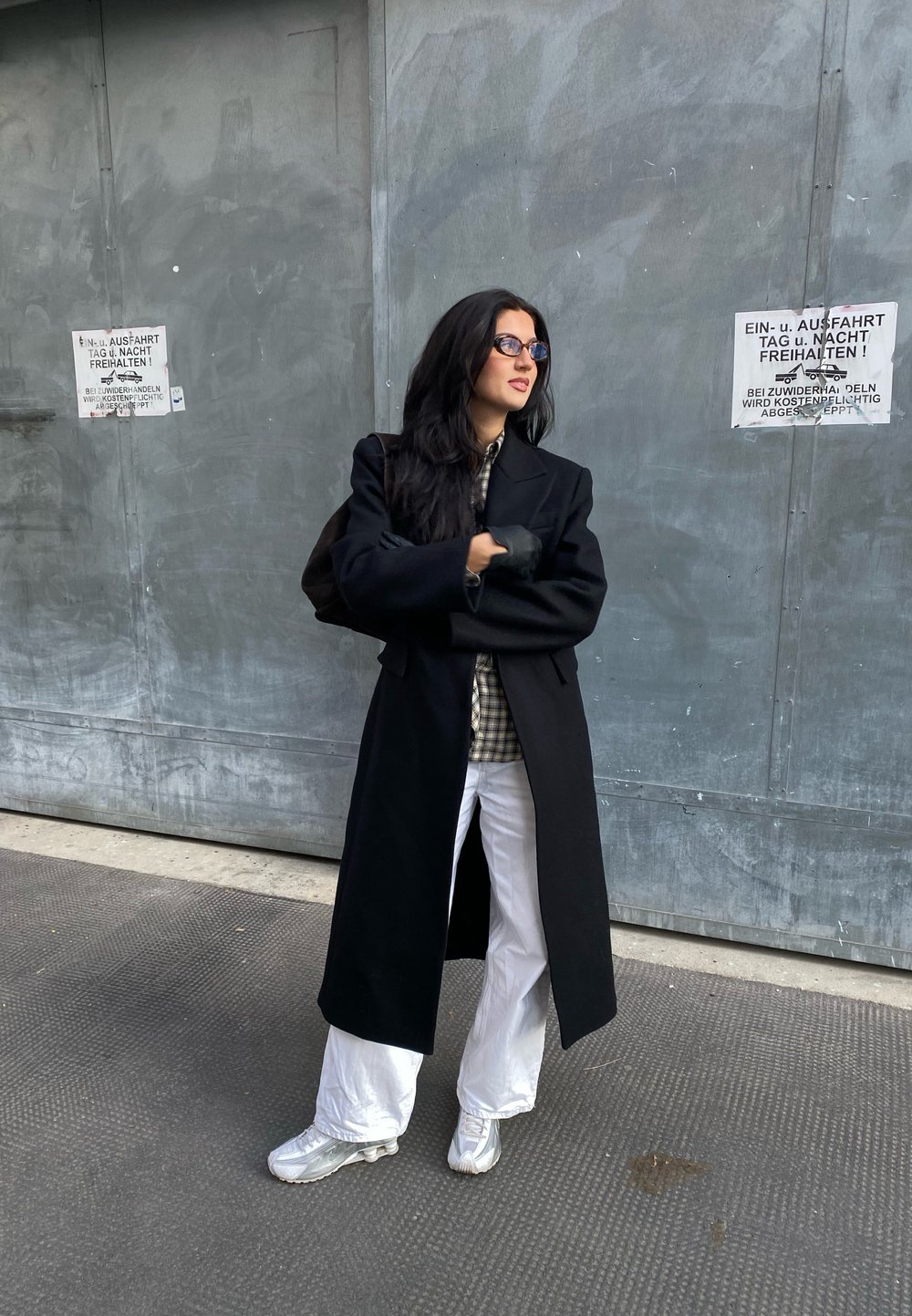 Μαύρο μακρύ παλτό πάνω από καρό πουκάμισο, συνδυασμένο με λευκά παντελόνια και ασημί αθλητικά παπούτσια. Στάση όρθια με τα χέρια σταυρωμένα μπροστά σε γκρι τοίχο.
