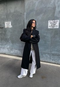 Μαύρο μακρύ παλτό πάνω από καρό πουκάμισο, συνδυασμένο με λευκά παντελόνια και ασημί αθλητικά παπούτσια. Στάση όρθια με τα χέρια σταυρωμένα μπροστά σε γκρι τοίχο.