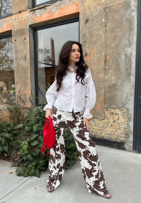 Weiße Bluse mit Knopfleiste und Puffärmeln; braun-weiße gemusterte weite Hose; rote Handtasche; strukturierter Hintergrund mit Grünpflanzen.