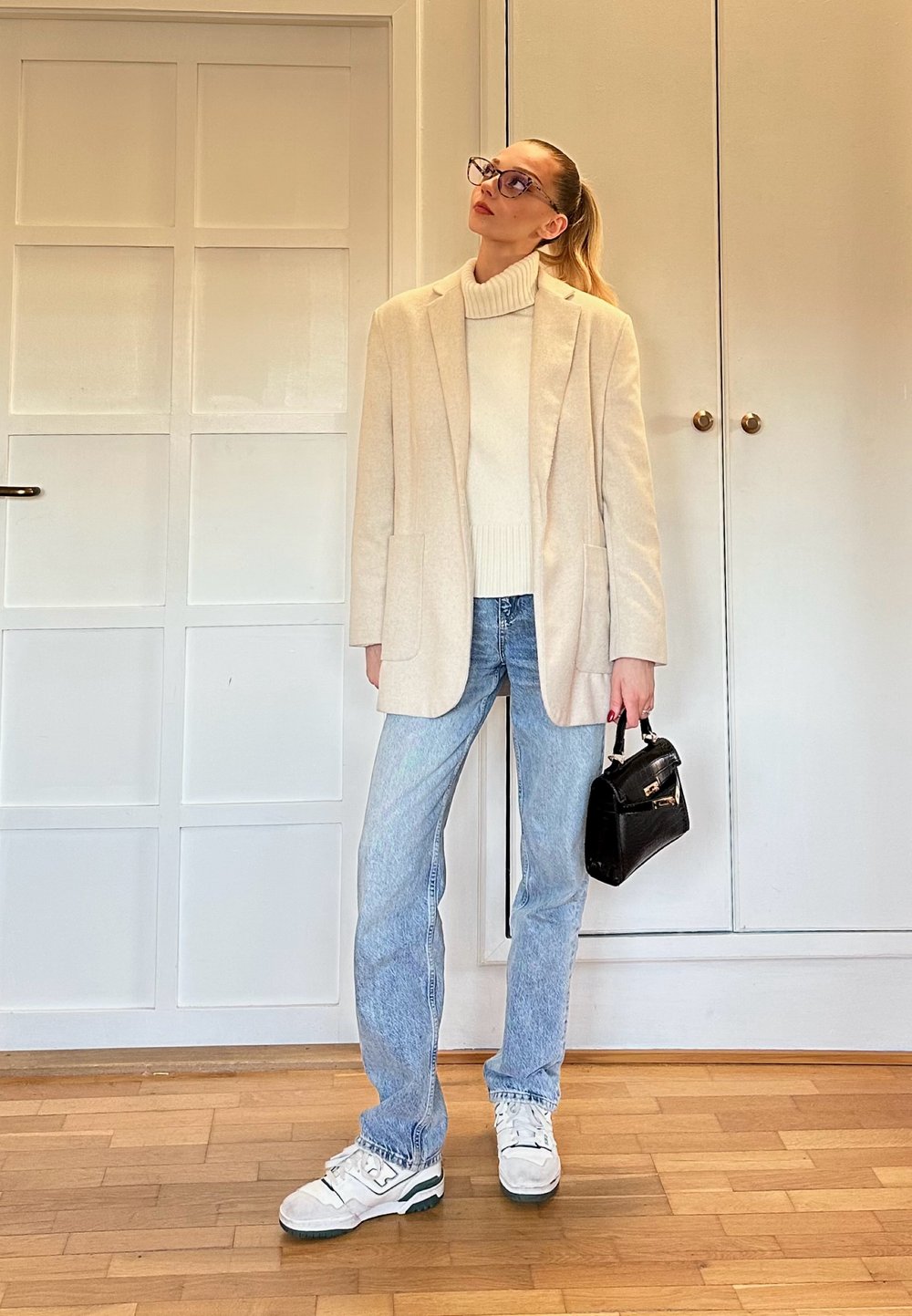 Blazer crème sur un col roulé blanc, jean bleu clair et baskets blanches avec des accents verts. Petit sac à main noir à la main.