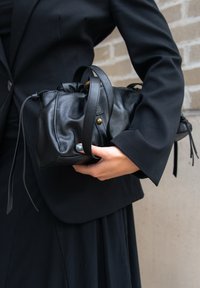 Oseba v črnem blazerju drži mehko črno usnjeno torbico z dolgo naramnico in podrobnostmi iz mesečnika proti ozadju zidov iz opeke.