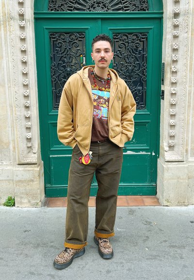 Sudadera con capucha marrón sobre una camiseta gráfica, pantalones verde oliva y botas de print de leopardo. Incluye un collar de cuentas y un llavero.