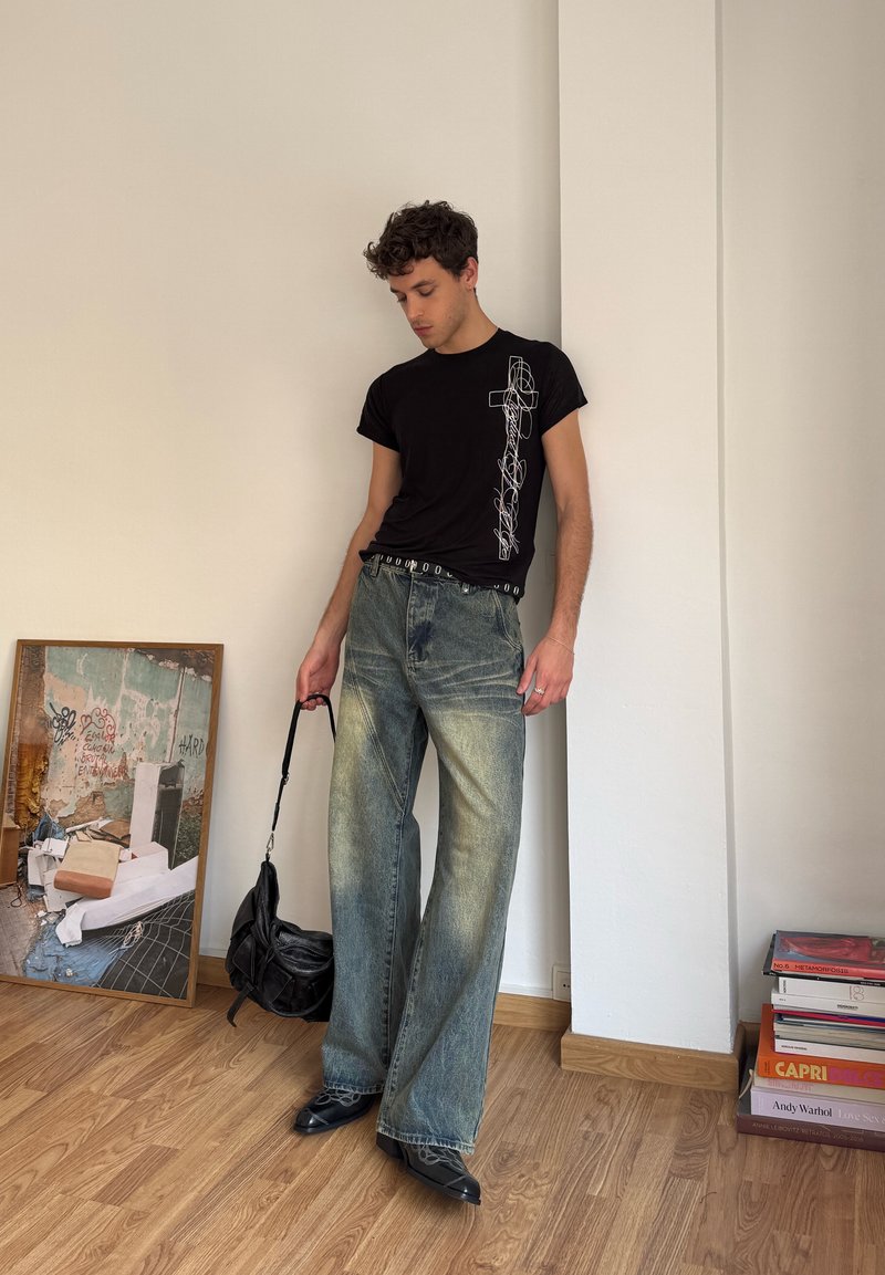 T-shirt nero con design grafico bianco, jeans larghi azzurri chiari, borsa nera e scarpe nere a punta, il tutto su un pavimento in legno contro un muro.