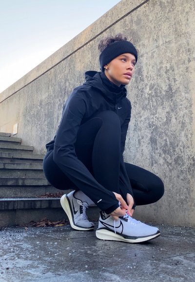 Chaqueta deportiva negra con capucha, leggins negros y zapatillas de running blancas. La persona está agachada en unos escalones de piedra, atándose los cordones.