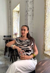 Zebra-gestreepte top met een ronde hals, witte broek en meerdere armbanden. Helder glas met een rietje in de hand, zittend in een modern café.