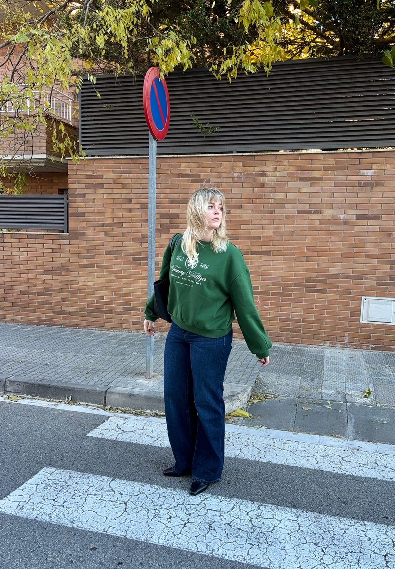 Femme en sweat-shirt vert et jean, debout sur un passage pour piétons près d'un panneau d'interdiction de stationner et d'un mur en briques, avec des feuilles d'automne au-dessus.