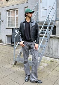 Czarna skórzana kurtka, czarny t-shirt, szare dzwony, czarne buty i zielona czapka. Stoi obok metalowych schodów w ceglanej scenerii.