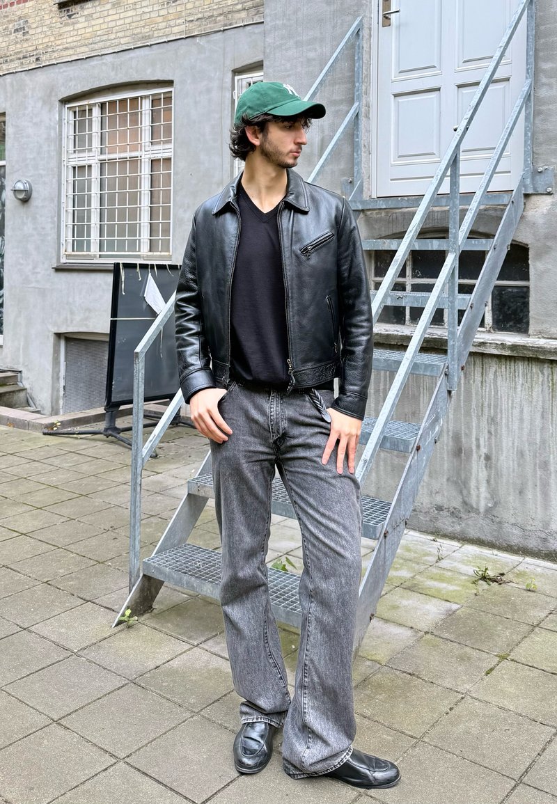 Mustast nahast jakk, must t-särk, hallid lehvitatud teksad, mustad kingad ja roheline nokamüts. Seisab metalltreppide kõrval tellisseinas.