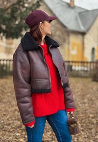 Jachetă maro scurtă din piele cu guler negru din blană sintetică, asortată cu un pulover roșu tricotat în model cu cabluri și blugi albaștri, ținând o poșetă mică maro.