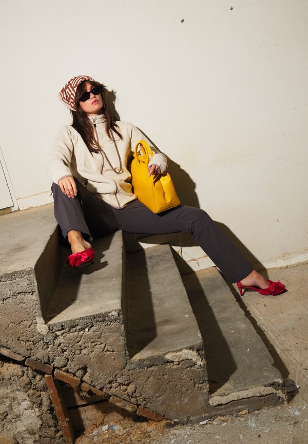 Mujer con gafas de sol, chaqueta beige y pañuelo estampado en la cabeza, sentada en escalones de hormigón ásperos, sosteniendo un bolso amarillo brillante y con tacones rojos.