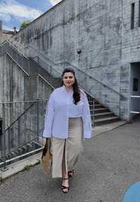 Camisa blanca de gran tamaño con detalles recortados en las mangas, falda midi beige con abertura frontal, sandalias negras de tiras y un bolso beige. Pared de hormigón de fondo.