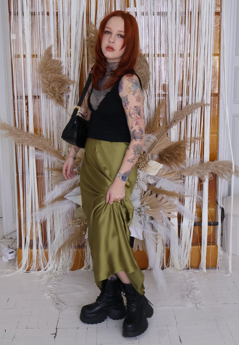 Jeune personne aux cheveux roux et tatouages portant un haut noir, une jupe en satin vert olive, des bottines compensées, posant devant une décoration florale séchée suspendue.