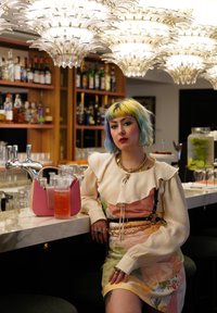 Ροζ τσάντα χεριού σε μάρμαρο μπαρ δίπλα σε ένα κοκτέιλ. Το μπαρ διαθέτει γυάλινα σκεύη και ράφια γεμάτα με διάφορες φιάλες. Μεγάλες κρυστάλλινες πολυέλαιοι από πάνω.