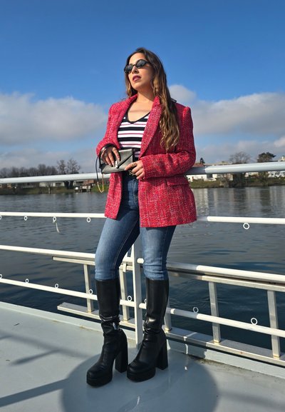 Blazer de tweed rojo sobre una blusa de rayas blancas y negras, jeans ajustados azules y botas altas de plataforma negras junto a una barandilla cerca del agua.