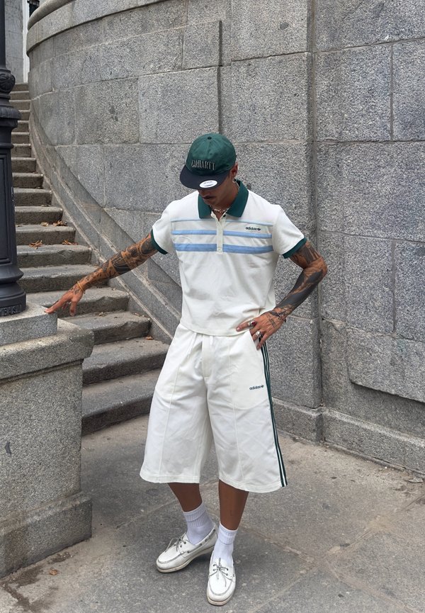 Polo bianco a maniche corte con colletto verde e strisce blu, abbinato a pantaloni corti bianchi larghi con strisce verdi laterali. Sfondo in pietra grigia.
