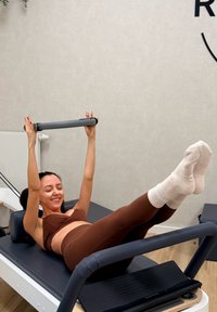 Fitnes reformer sivo platformo in oblazinjenim podporami, kjer oseba v rjavi športni opremi izvaja vajo z sivim krmilom.