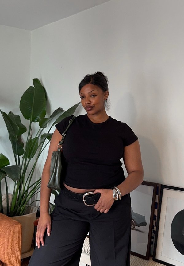 Frau in schwarzem T-Shirt und Hose posiert mit der Hand am Gürtel, trägt eine Umhängetasche, steht neben großen grünen Pflanzen und gerahmter Kunst im Raum.