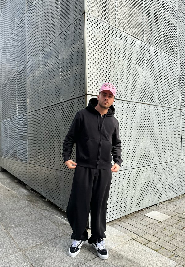 Sweatshirt preta com fecho na frente e bolsos, combinada com calças de fato pretas ajustadas. Usada com um boné rosa claro e sapatilhas pretas. Fundo texturizado.