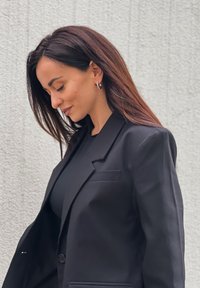 Czarna marynarka dopasowana z pojedynczym guzikiem, klapkami i wyraźnie zarysowanymi ramionami; noszona na czarnym topie, o gładkiej tkaninie i prostych liniach.