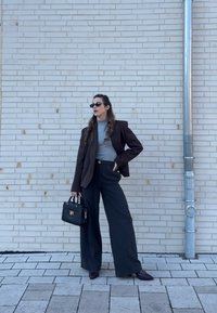 Γυναίκα που φοράει γυαλιά ηλίου, καφέ σακάκι, γκρι ζιβάγκο, μαύρο φαρδύ παντελόνι, κρατώντας μαύρη τσάντα, όρθια δίπλα σε φωτεινό τοίχο από τούβλα κοντά σε μεταλλικό σωλήνα.