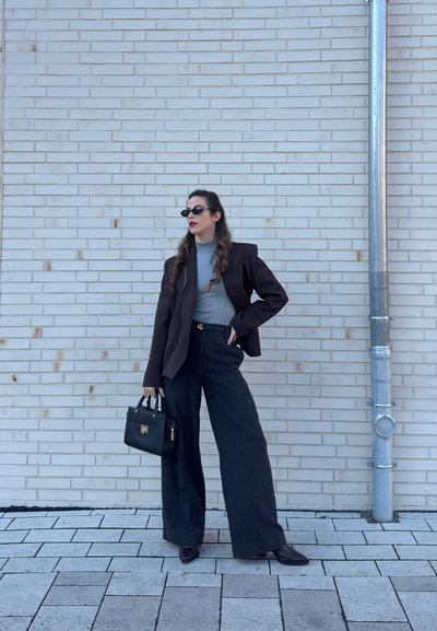 Mujer con gafas de sol, blazer marrón, camiseta de cuello alto gris, pantalones anchos negros, sosteniendo un bolso negro, de pie contra una pared de ladrillo claro cerca de una tubería de metal.