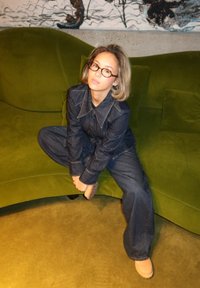 Τζιν ολόσωμη φόρμα με γιακά και μακριά μανίκια, με αντίθεση ραφής, φορεμένη με μπεζ παπούτσια σε έναν πράσινο καμπυλωτό καναπέ.