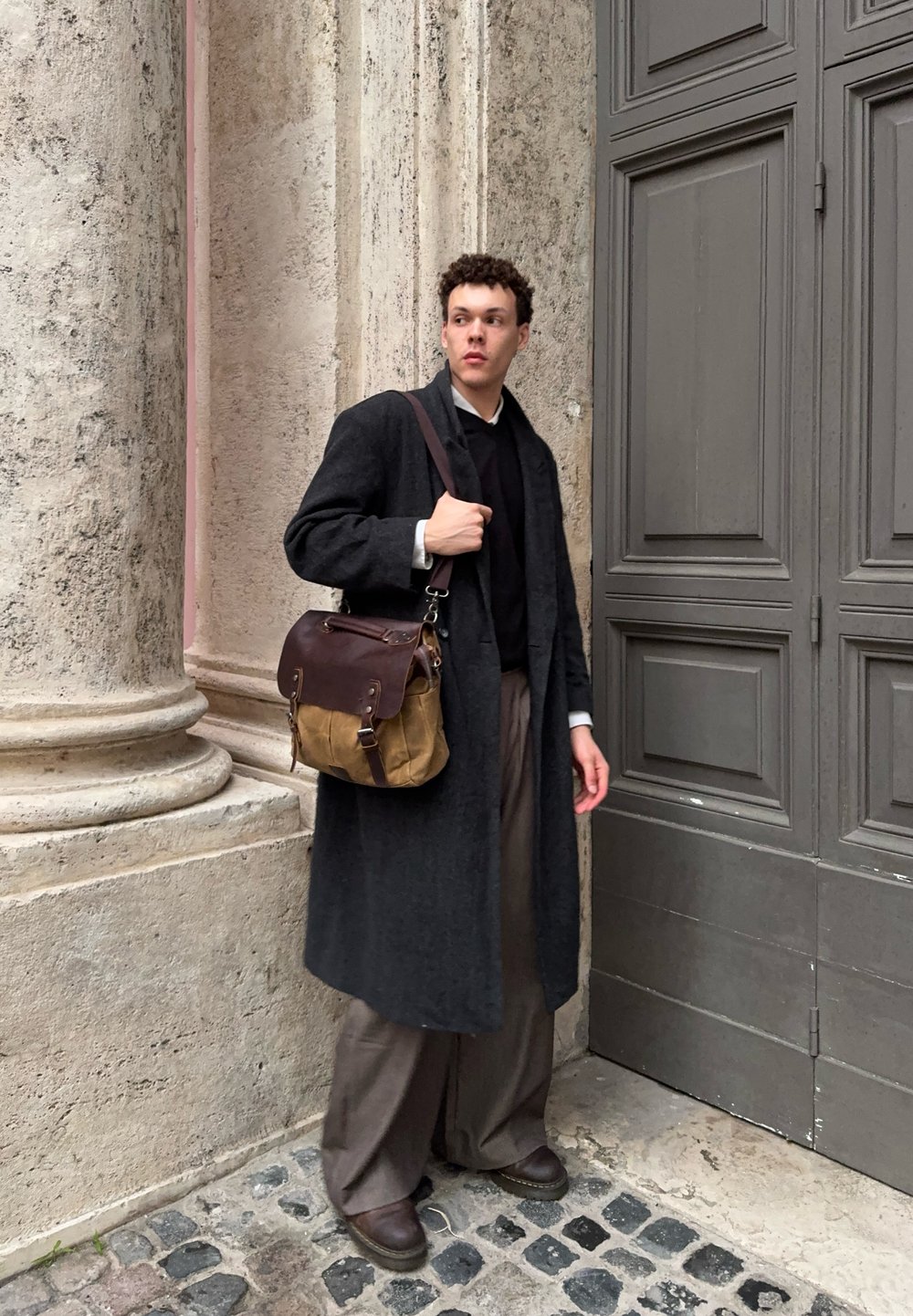 Tumšs mētelis, melns džemperis un plata brūna bikses. Brūna un pelēka soma ar metāla detalēm, nēsāta pāri plecam. Strukturēta akmens fona.